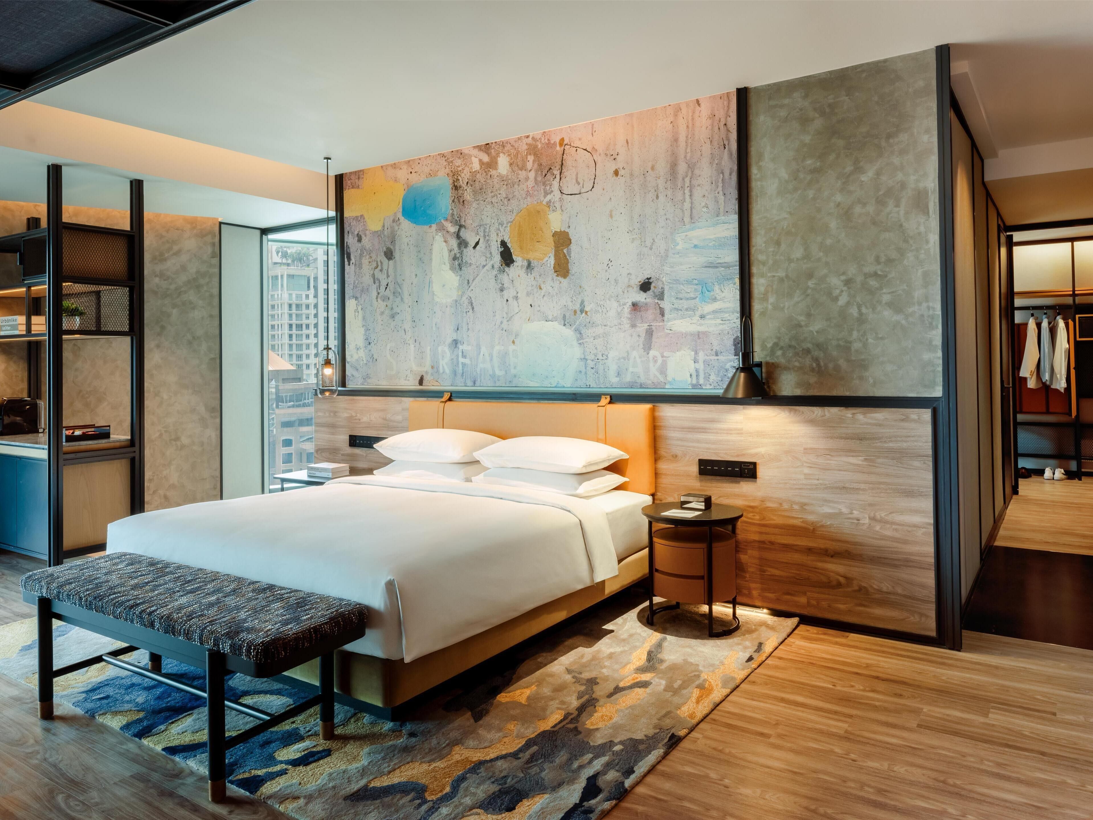 A modern hotel room with a large bed, a bench, a small table, and a large abstract painting on the wall.