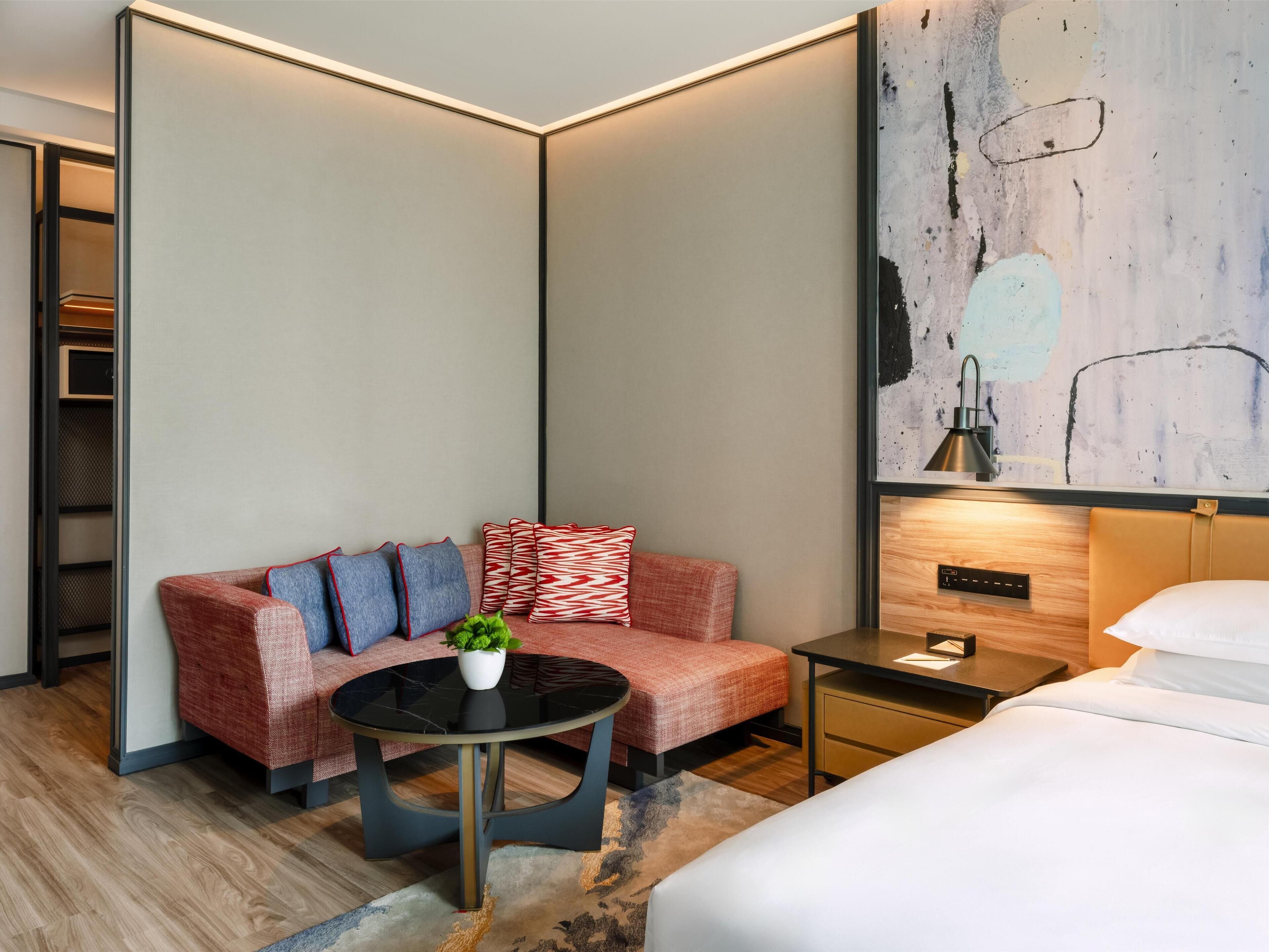 A modern hotel room with a sofa, a small round table with a plant, a bed, a nightstand, and a large abstract painting on the wall.