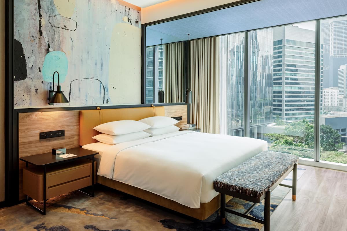 A modern hotel room with a large bed, a bench, a nightstand, a wall art piece, and a view of the city through large windows.