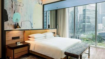 A modern hotel room with a large bed, a bench, a nightstand, a wall art piece, and a view of the city through large windows.