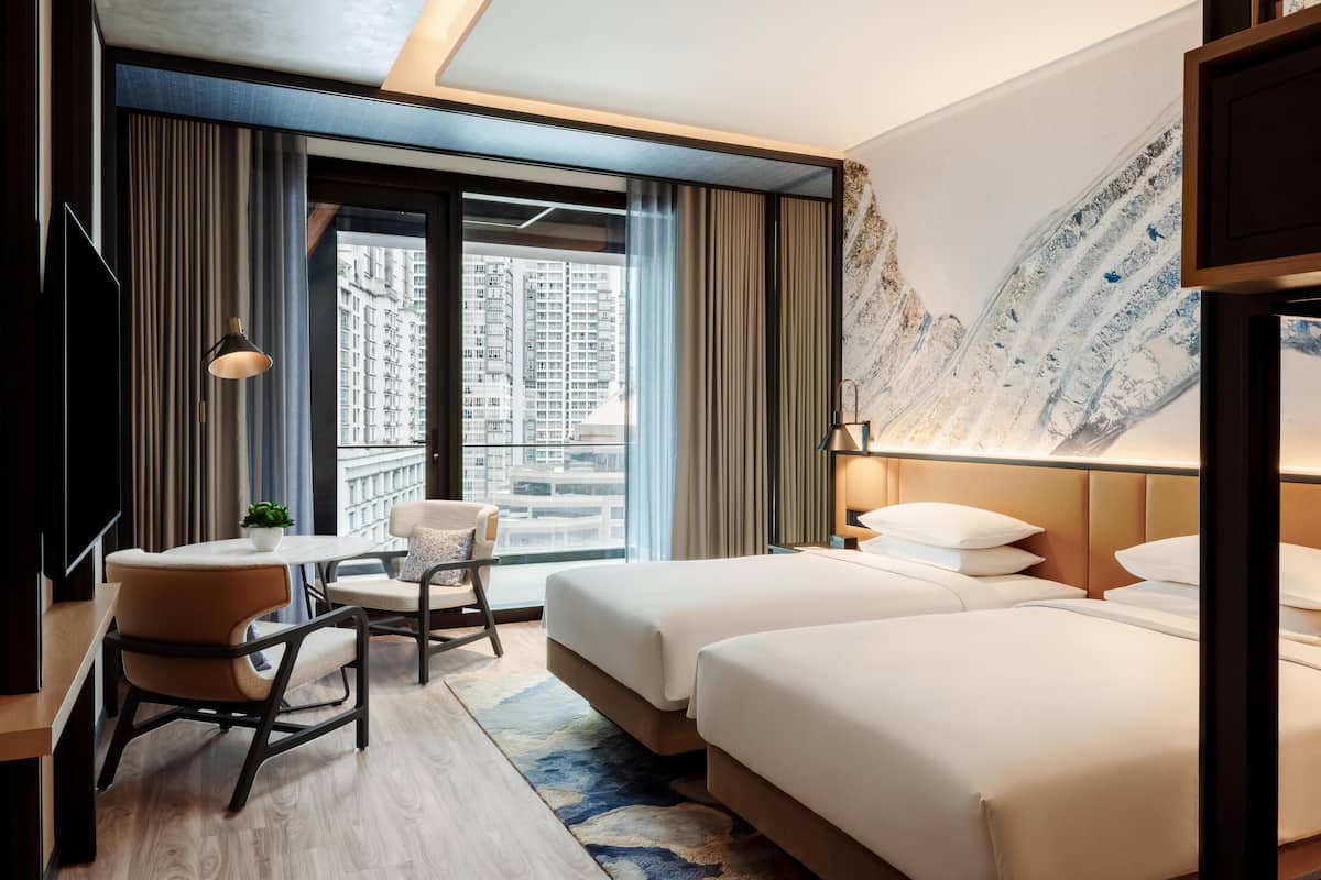 A hotel room with a large window, a desk with a chair, two beds, and a mounted TV.