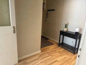 Um corredor com piso de madeira, uma porta branca, um console preto com uma planta e um abajur, e um gancho fixado na parede.