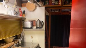 Kompakti keittiö, jossa on pesuallas, liesi ja erilaisia keittiövälineitä. Pieni jääkaappi on näkyvissä, ja työtasolla on puinen leikkuulauta.