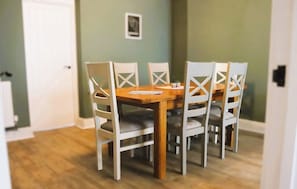 Un comedor con mesa de madera y sillas blancas, una puerta al fondo y un cuadro en la pared.