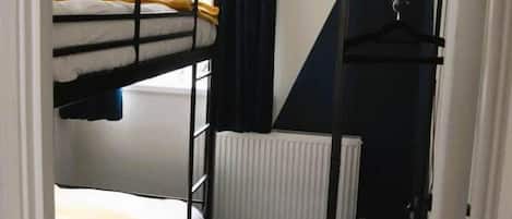 Ein schmaler Raum mit Etagenbetten, einem Heizkörper, einer Pendelleuchte und einem kleinen Holzregal mit zusammengeklappten Handtüchern.