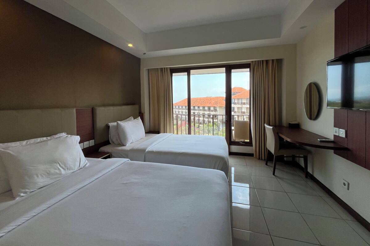 Habitación de hotel con dos camas, televisión, escritorio y balcón con vista a edificios.