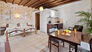 A room with a bed, a dining table with chairs, a kitchen area, and a stone wall. - Inia Lara Suites by Nomads (Paphos)