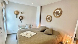 A bedroom with a large bed, two wall-mounted decorative items, a bedside table with a lamp, and a wicker chair. - Clara Beach Blue Infinity (Cumbre del Sol)