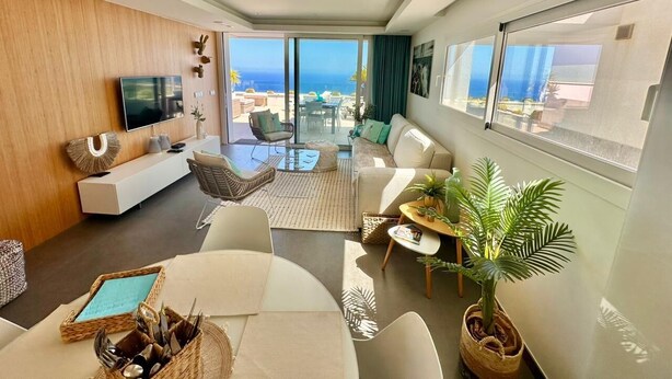 A modern living room with a large TV, comfortable seating, a dining area, and a balcony with a view of the ocean. - Clara Beach Blue Infinity (Cumbre del Sol)