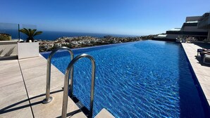 A swimming pool with metal railings, a clear blue sky, and a view of a coastal town. - Clara Beach Blue Infinity (Cumbre del Sol)