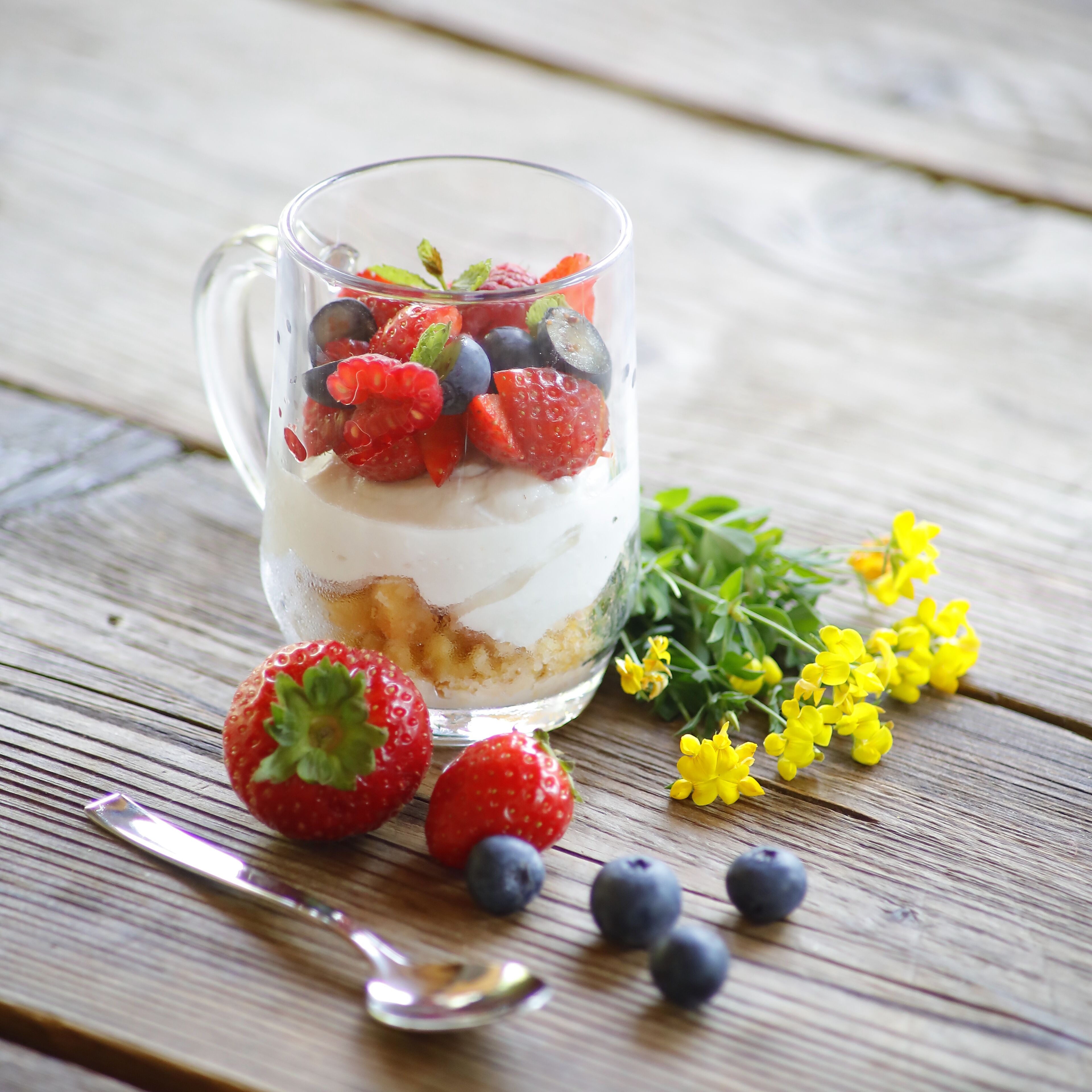 A layered dessert in a clear glass mug with strawberries, blueberries, and a creamy base, placed on a wooden surface with a spoon and fresh strawberries and blueberries nearby.