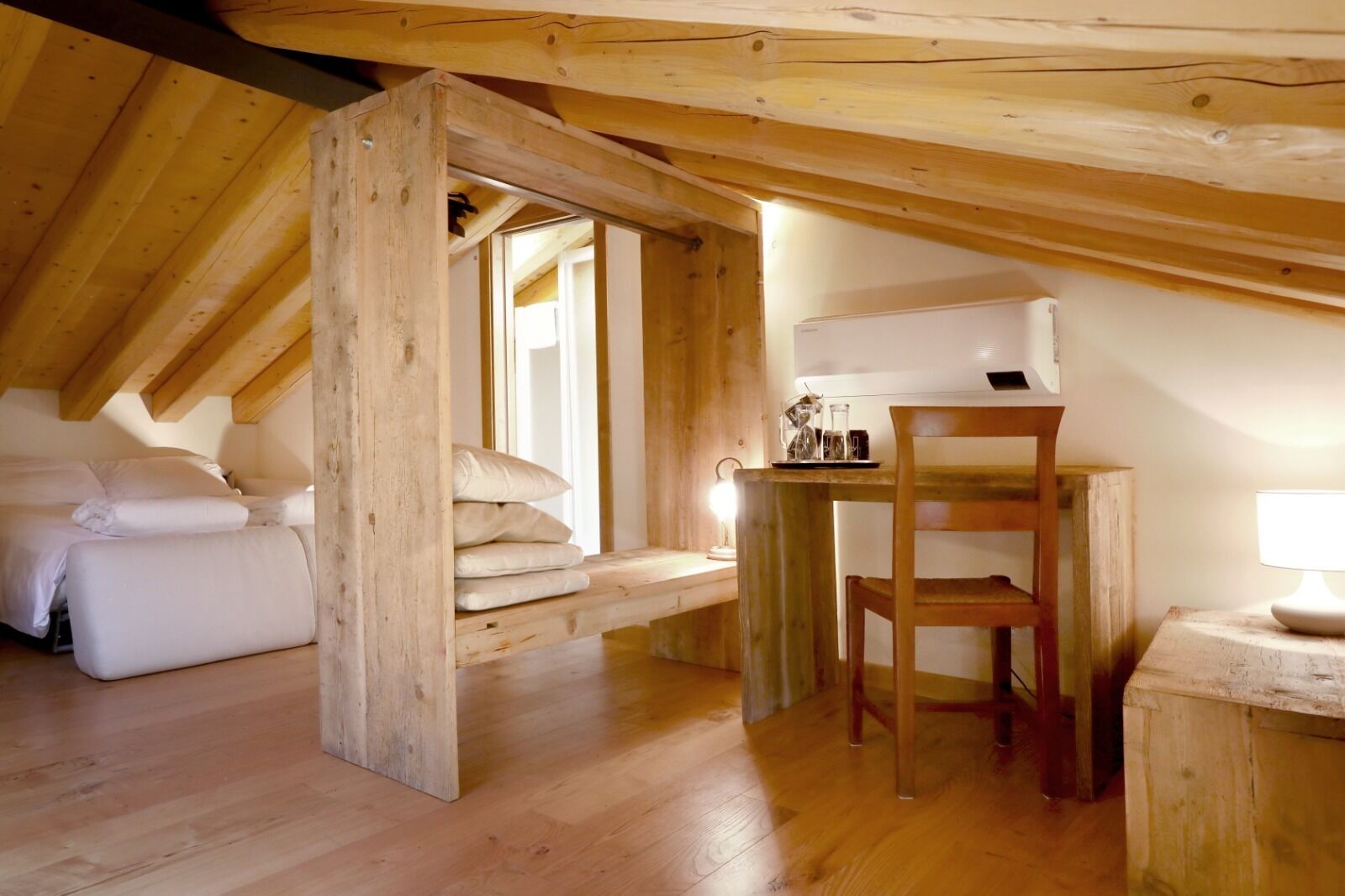 A cozy attic bedroom with a wooden ceiling, a bed, a small table with a chair, and a lamp.