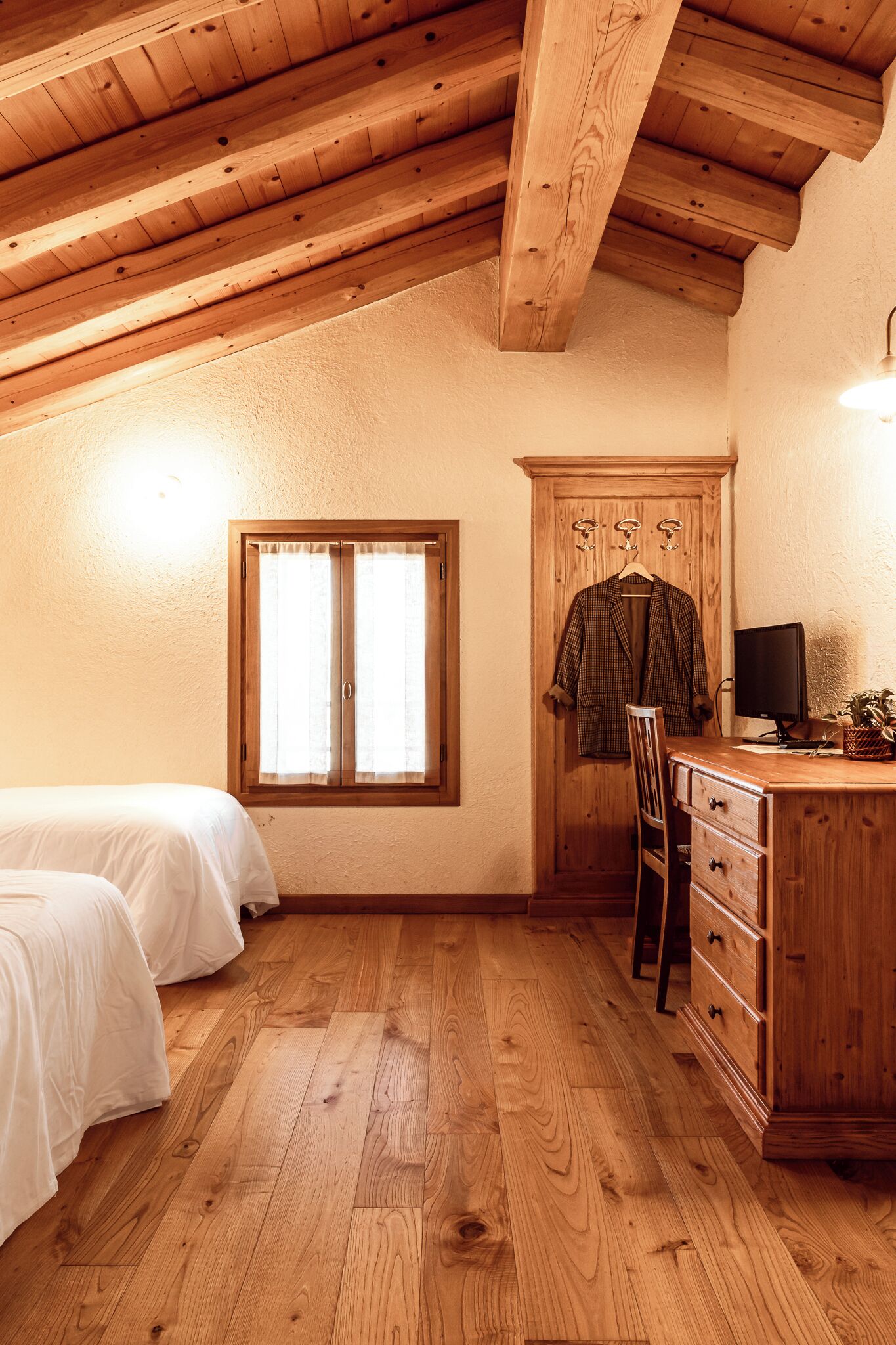Un dormitorio con piso de madera, una cama, un escritorio con silla, un armario y una ventana con cortinas.