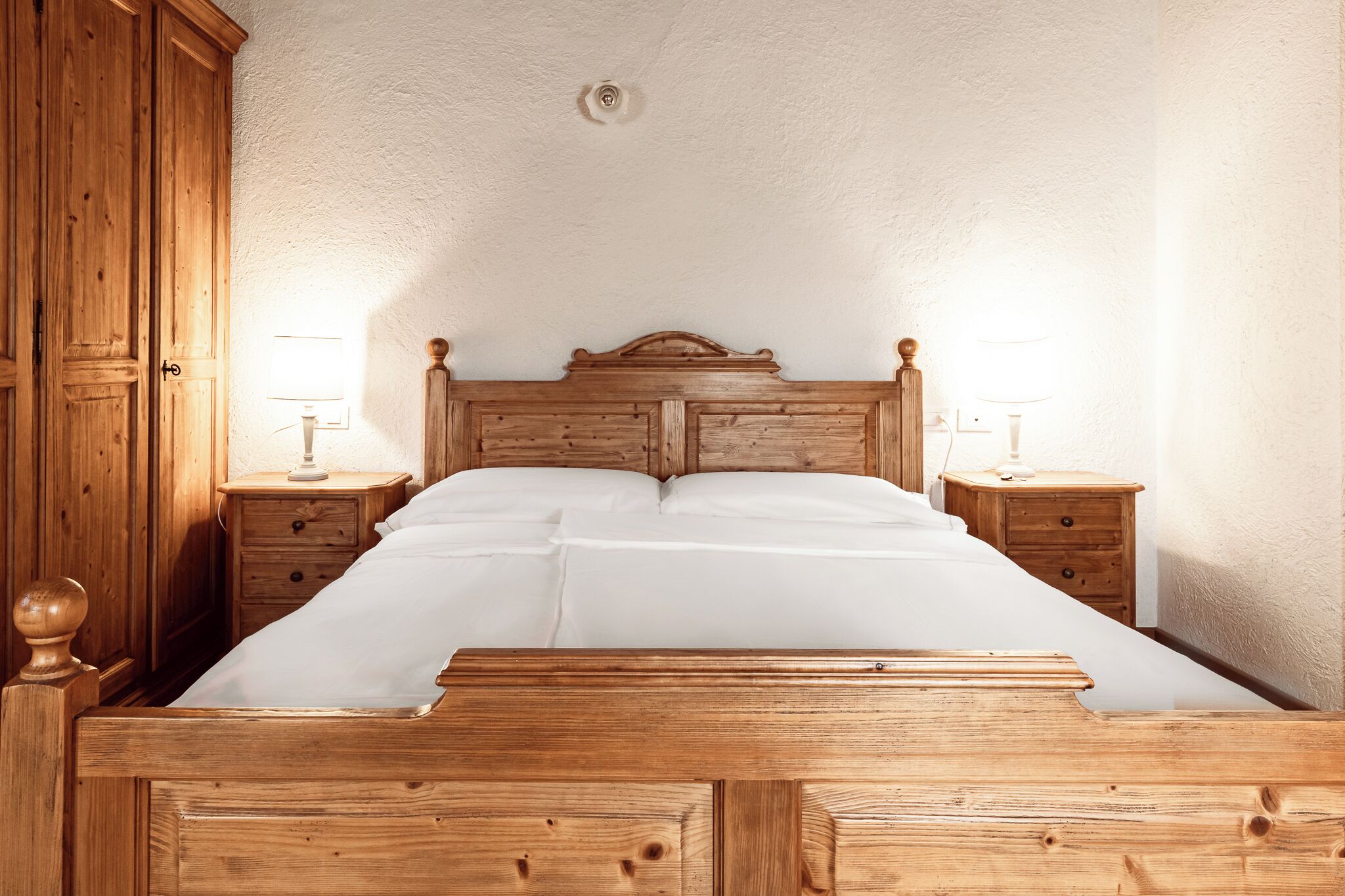 A wooden bed with white linens, two bedside tables with lamps, and a wooden wardrobe.