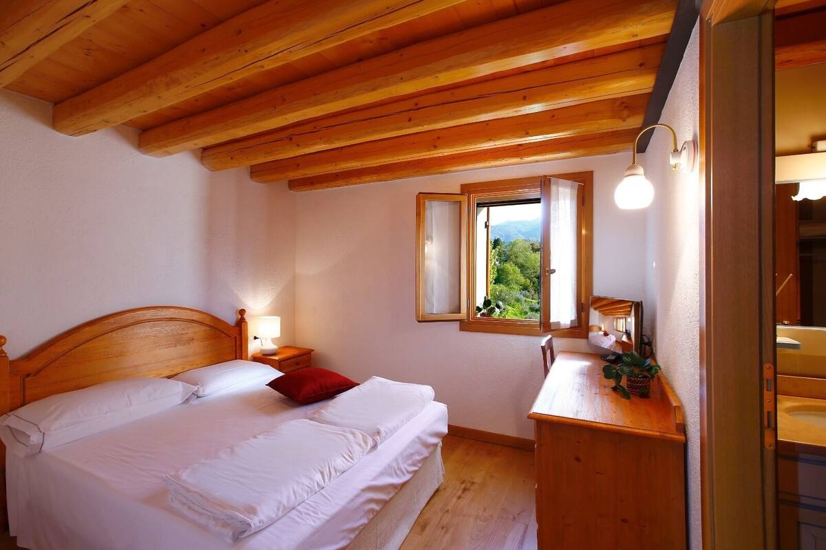 Un dormitorio con techo de madera, una cama con ropa blanca, una mesita de noche con lámpara, un ventanal con vistas a la vegetación y un aparador de madera.