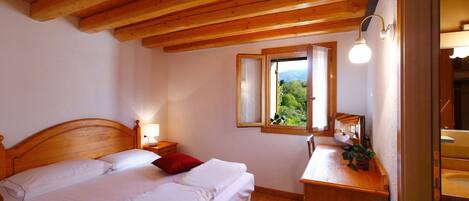 Un dormitorio con techo de madera, una cama con ropa blanca, una mesita de noche con lámpara, un ventanal con vistas a la vegetación y un aparador de madera.
