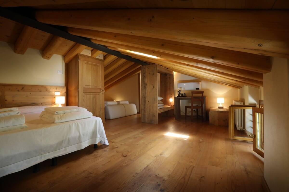 Un dormitorio de buhardilla acogedor con techo de madera, cama con ropa blanca, escritorio con silla y una mesita de noche con lámpara.