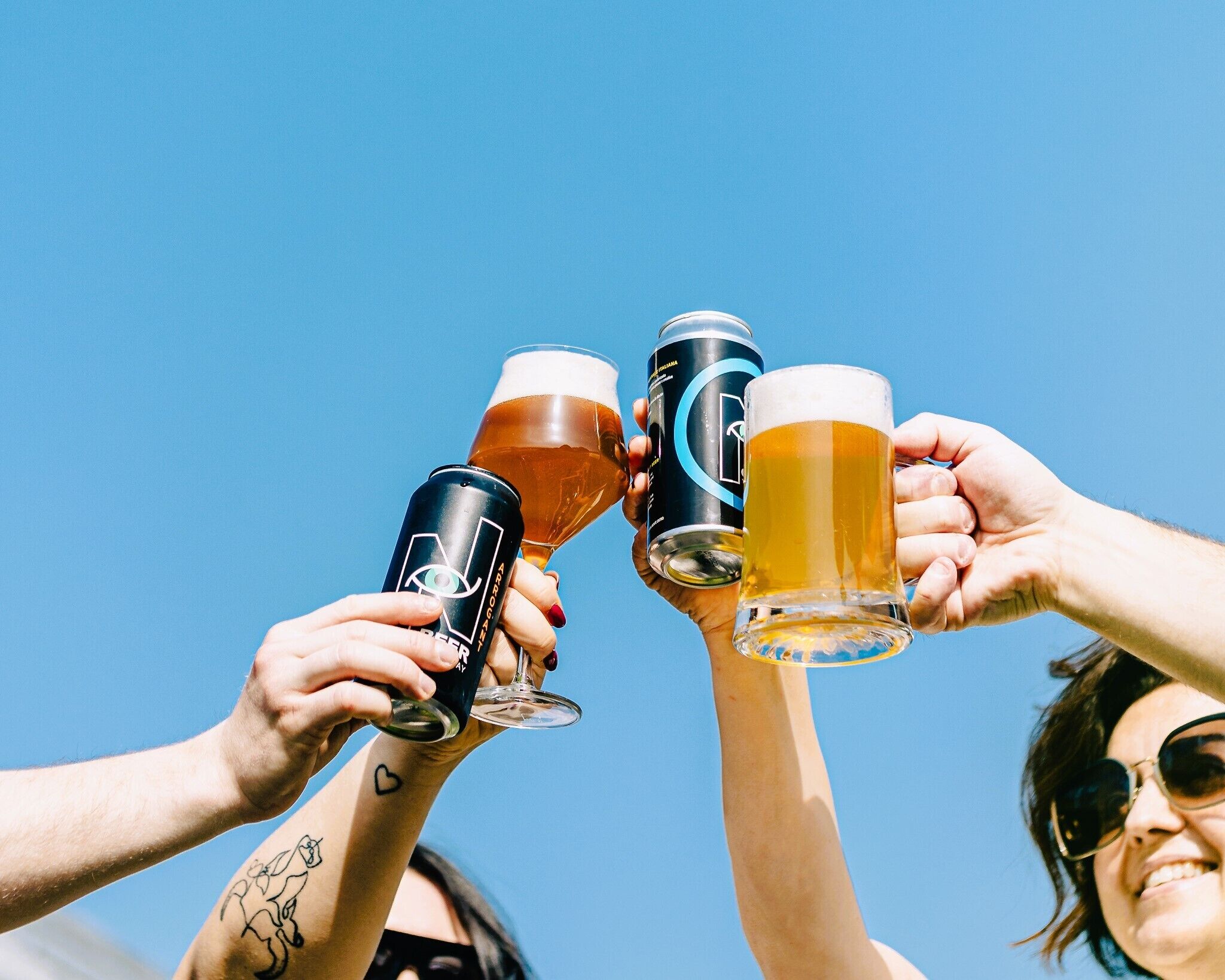 Dos personas brindando con vasos de cerveza y una lata, frente a un cielo azul despejado.