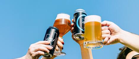 Dos personas brindando con vasos de cerveza y una lata, frente a un cielo azul despejado.