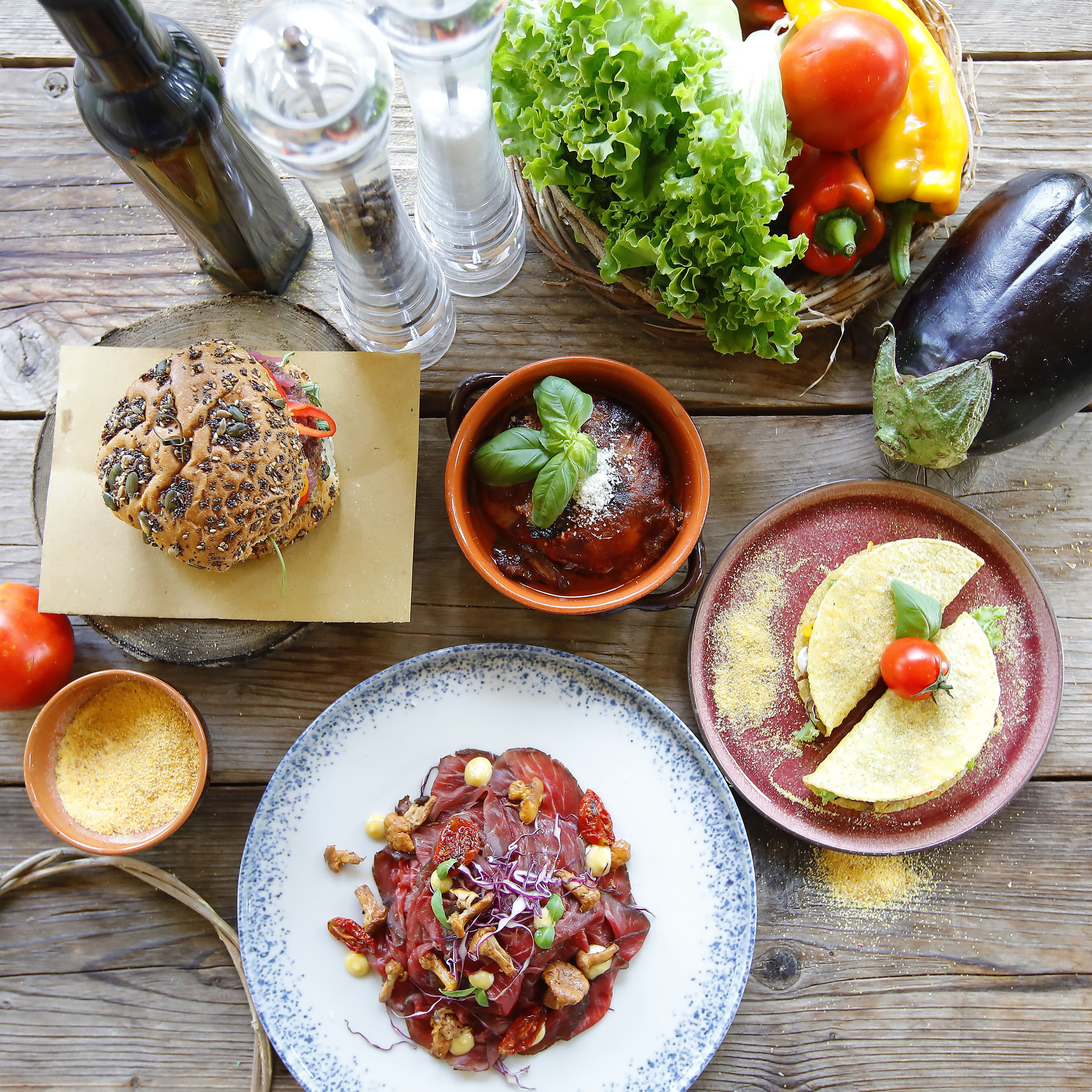 Ein Holztisch mit verschiedenen Speisen, darunter ein Burger, eine Suppenschüssel und eine Fleischplatte.