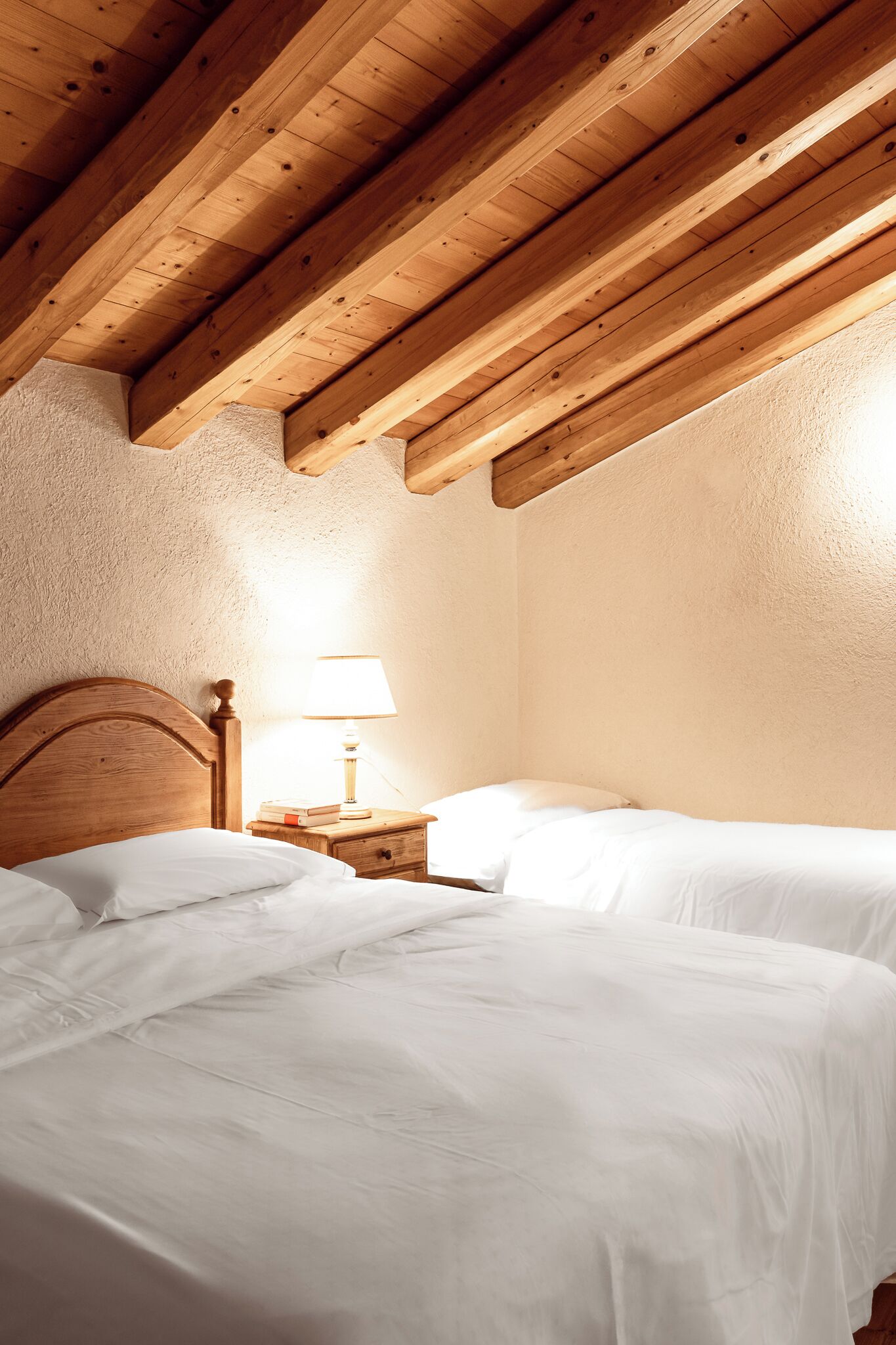 Een slaapkamer met twee bedden, houten balken in het plafond, een nachtkastje met een lamp en een gestructureerde wand.
