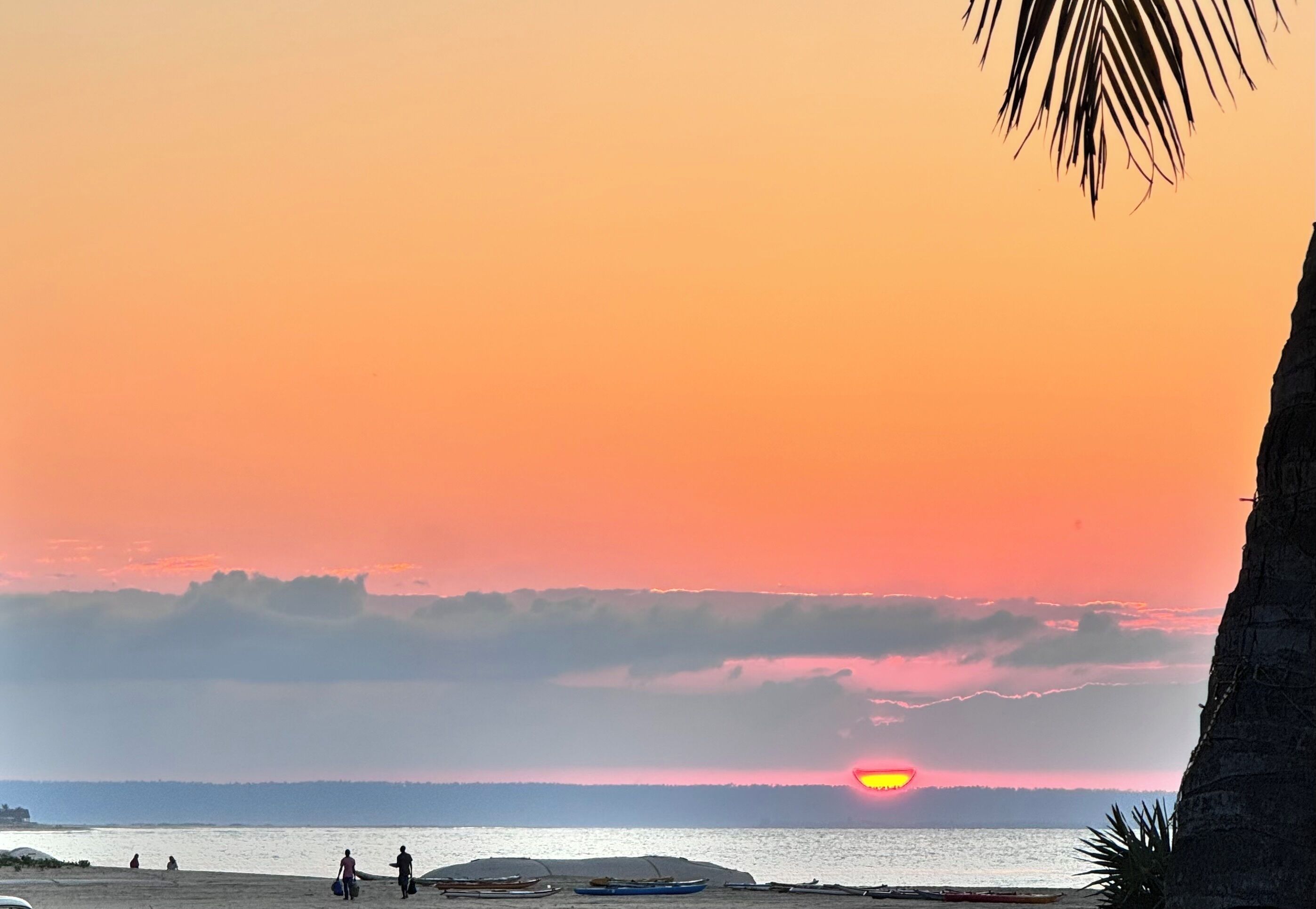 Escena de playa con atardecer, barcos y gente.