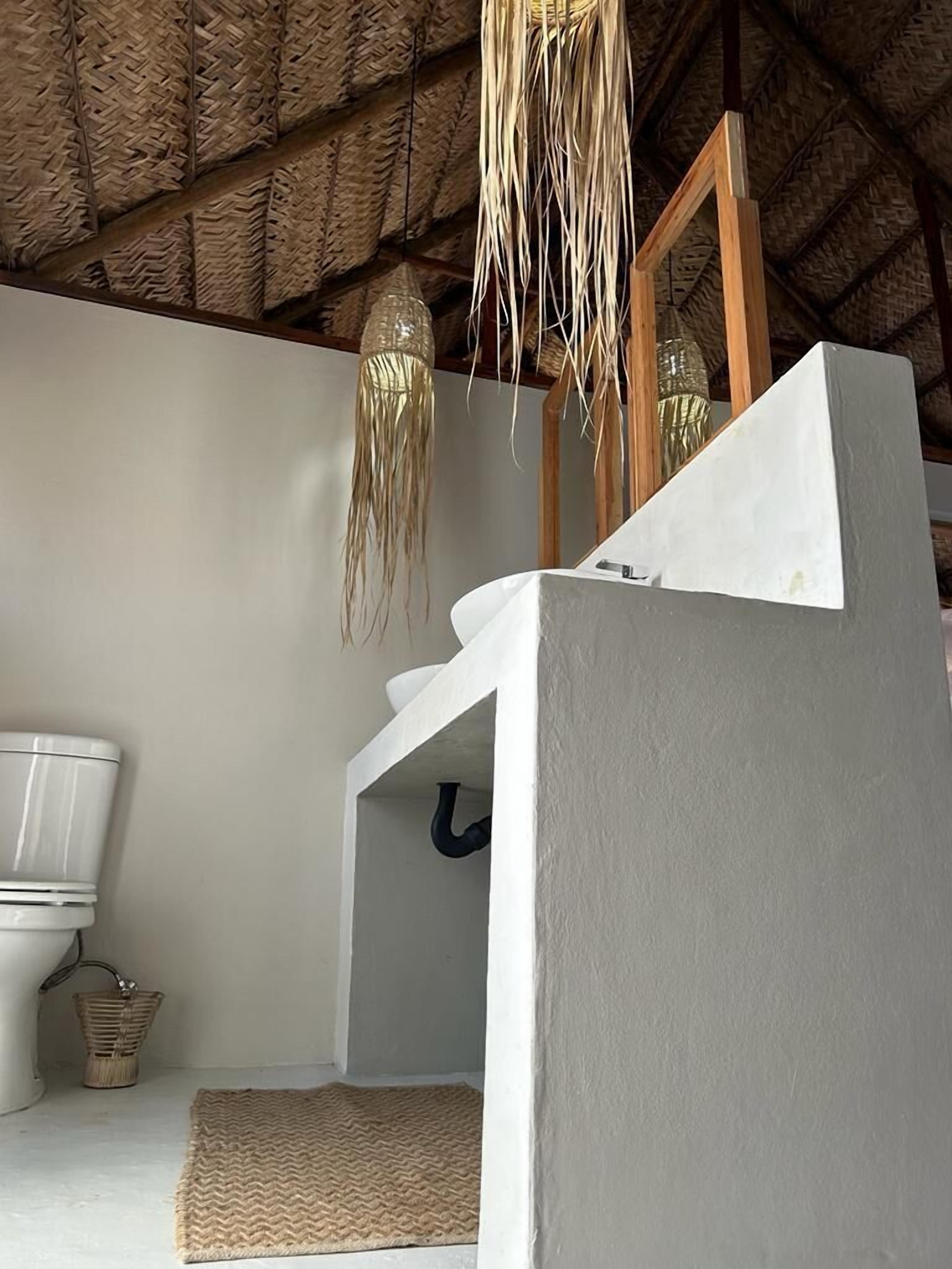 Un baño con inodoro blanco, un tapete tejido en el suelo y una lámpara colgante.