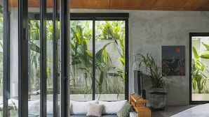 Een moderne woonkamer met een grote glazen deur die uitkomt op een balkon, een bank met kussens, een klein houten tafeltje en een plant in pot.