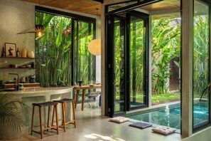 A modern kitchen with a bar area, large windows, and a view of a lush garden and pool.