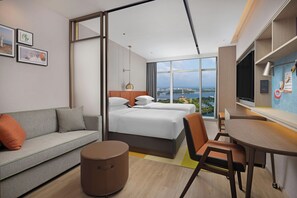 A modern hotel room with a bed, a sofa, a dining area, and a view of a cityscape.