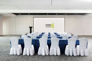 Un salón de conferencias con mesas dispuestas para una reunión, sillas blancas y una pantalla que muestra la marca 'HOME 2'.