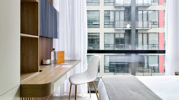 Una habitación de hotel moderna con un escritorio de madera, una silla blanca, una cama con manta estampada y un ventanal con vistas a la ciudad.