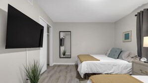 A bedroom with two beds, a TV mounted on the wall, a bedside table with a lamp, a potted plant, and a mirror.