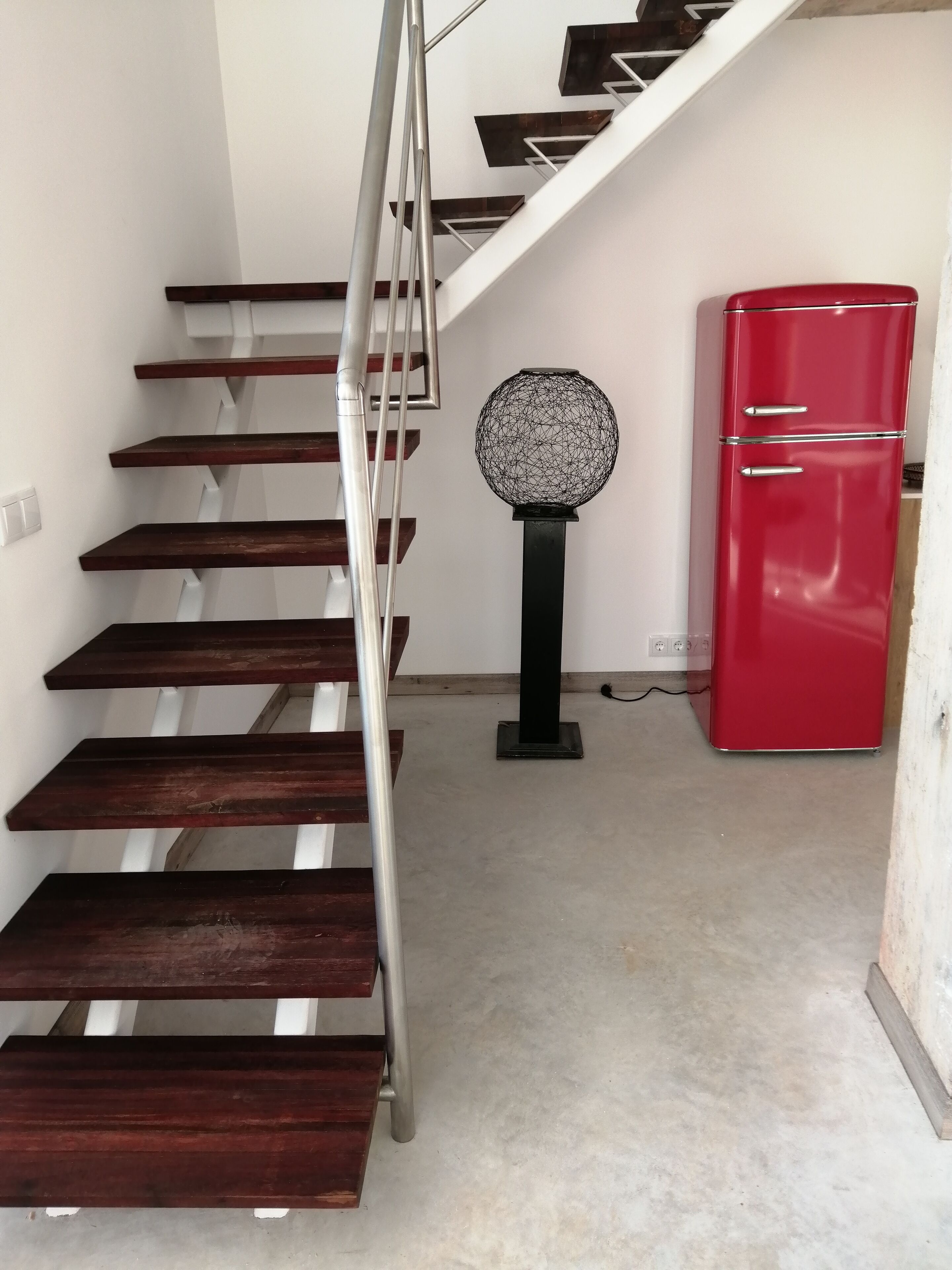 Eine Treppe mit Holzbodenstufen und Metallgeländer, ein roter Kühlschrank und ein dekorativer, kugelförmiger Gegenstand auf dem Boden.
