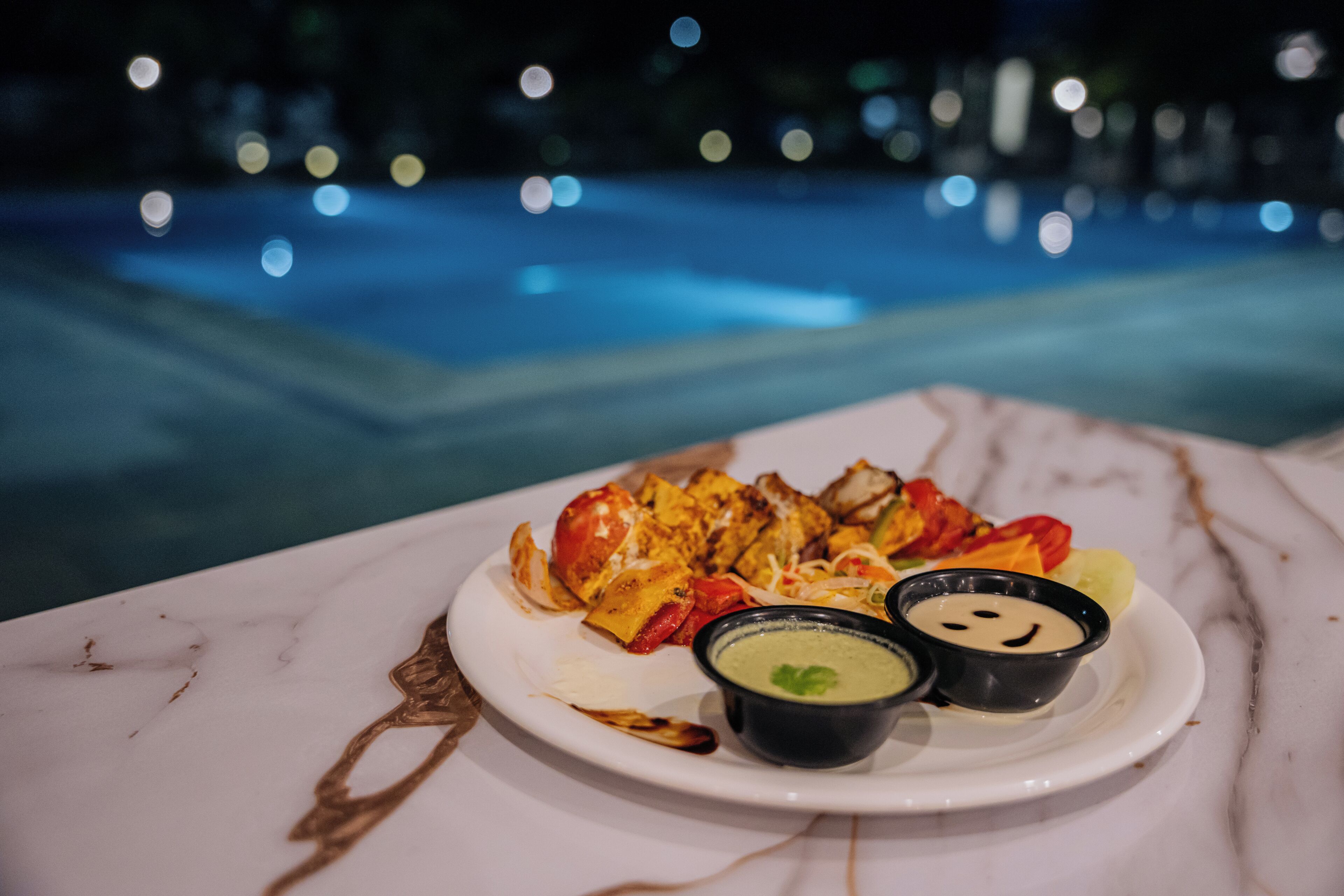 A plate of food with two dipping sauces on a marble table near a pool.