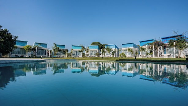 Um hotel moderno com uma piscina refletora na frente, apresentando uma série de edifícios com telhados azuis e palmeiras.