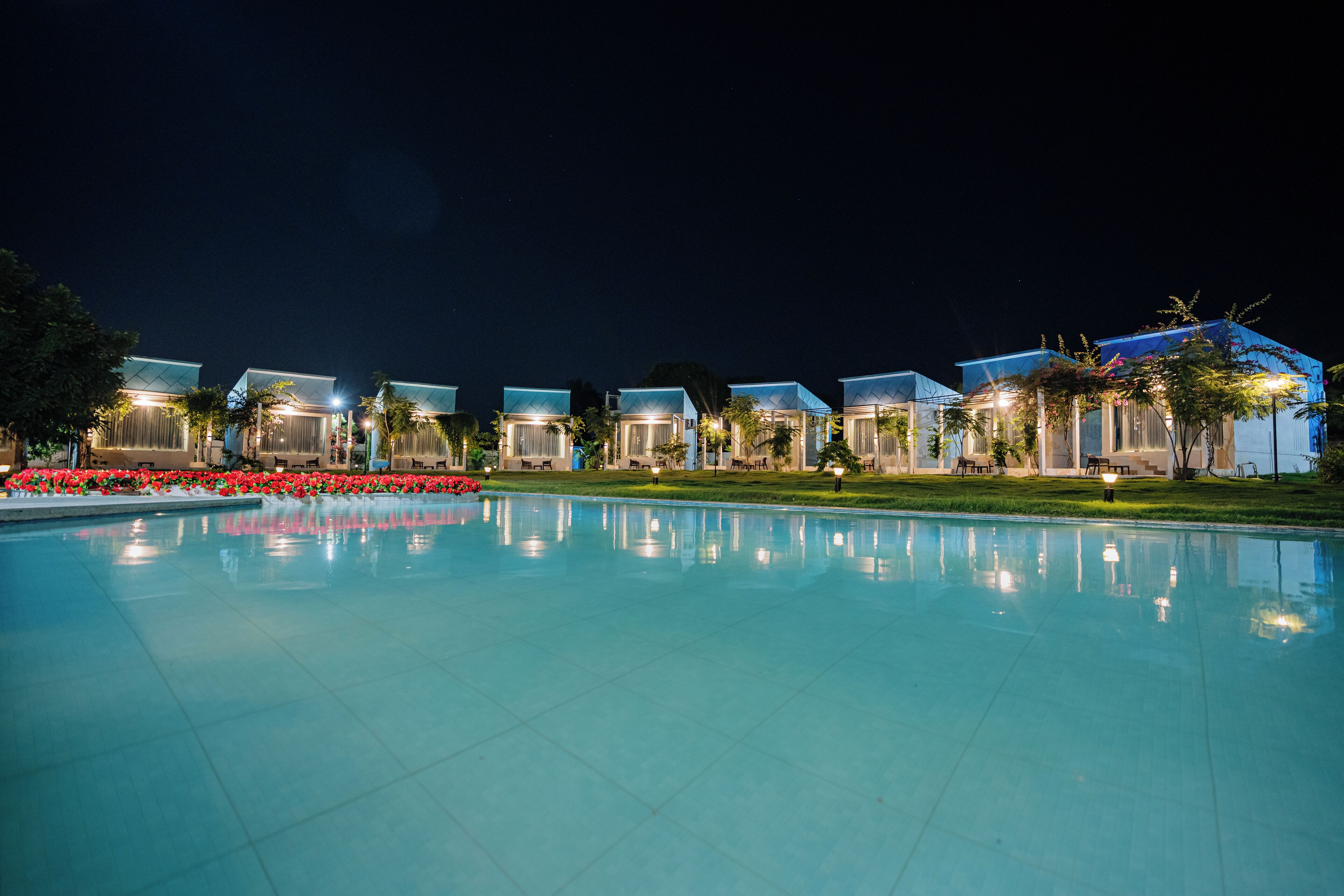 A nighttime view of a hotel with a swimming pool, multiple guest rooms, and a well-lit garden area.