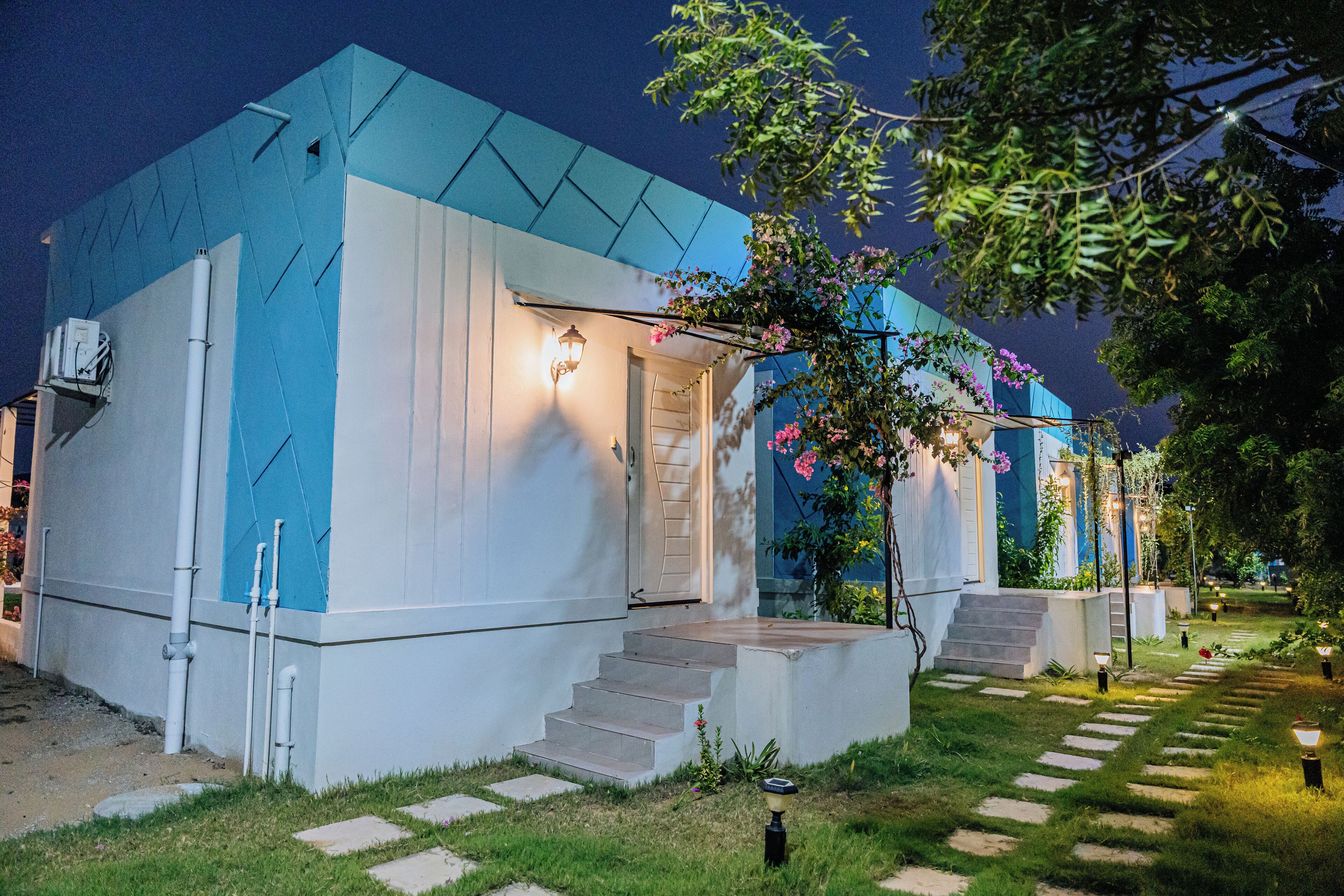 A modern building with a blue and white facade, a staircase leading to the entrance, and a garden area with stepping stones and lights.