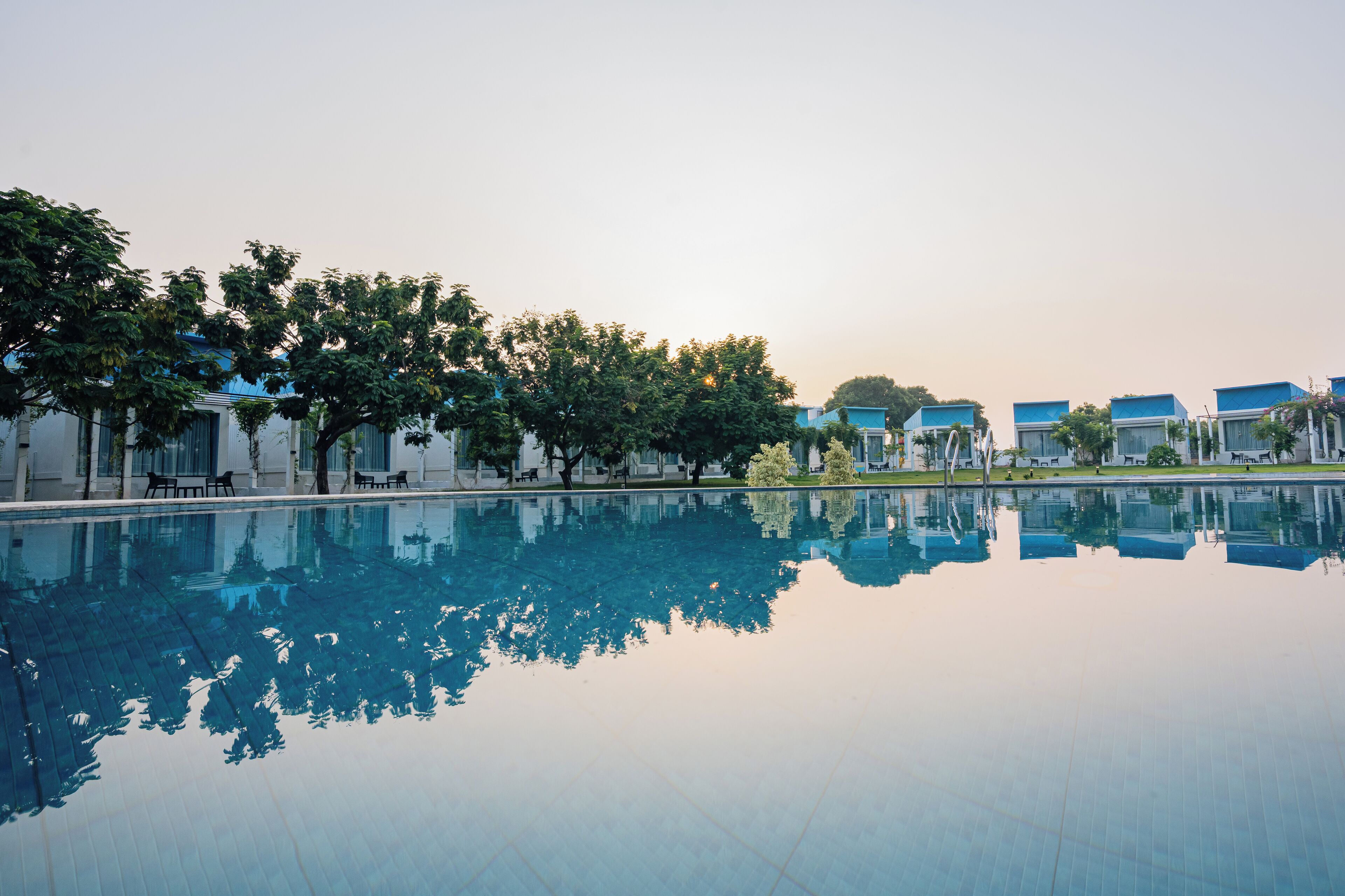 A serene swimming pool with clear reflections, surrounded by trees and buildings with blue roofs.