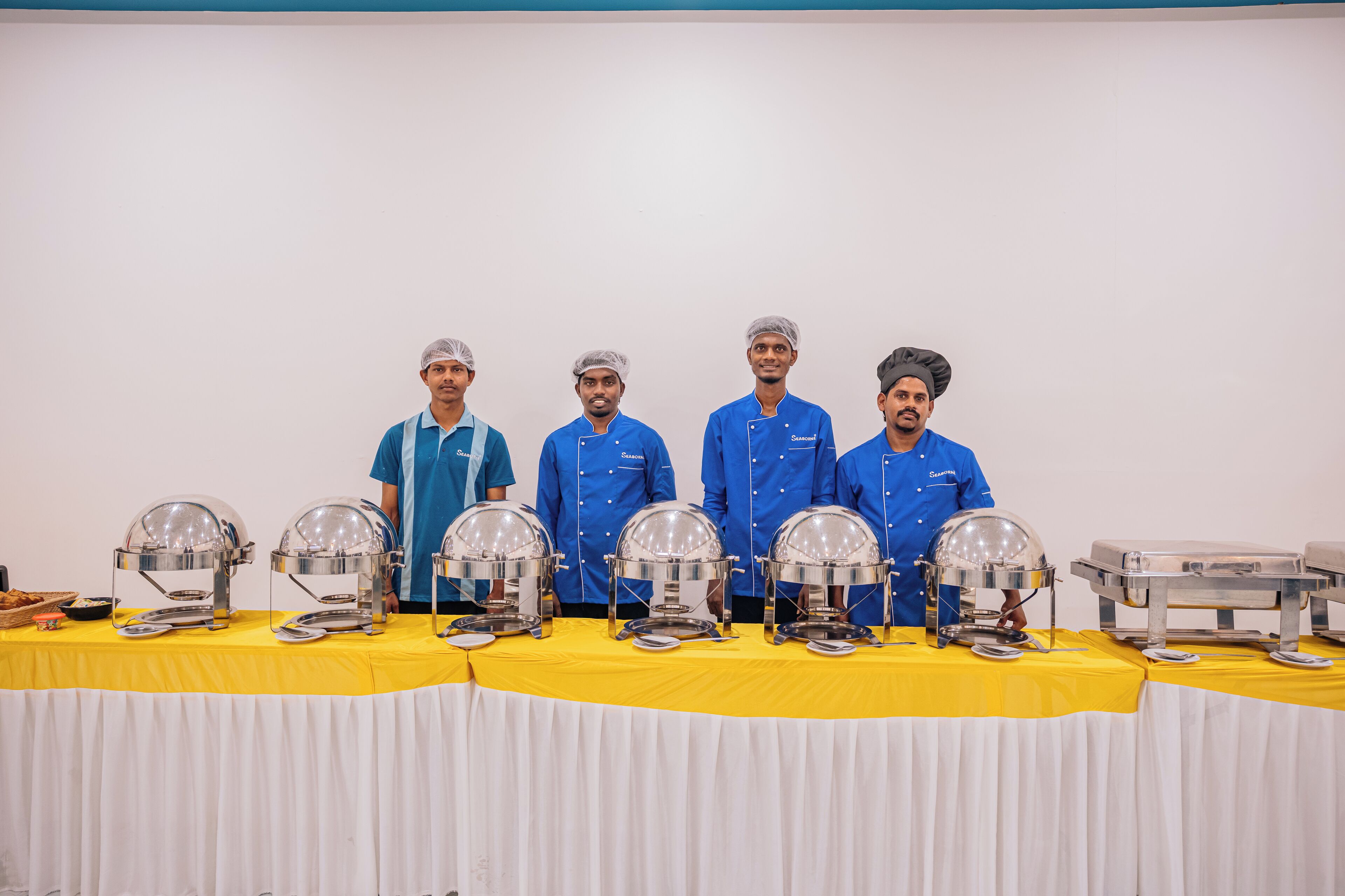 Four individuals in blue uniforms standing behind buffet service stations with clear dome covers and silver trays.