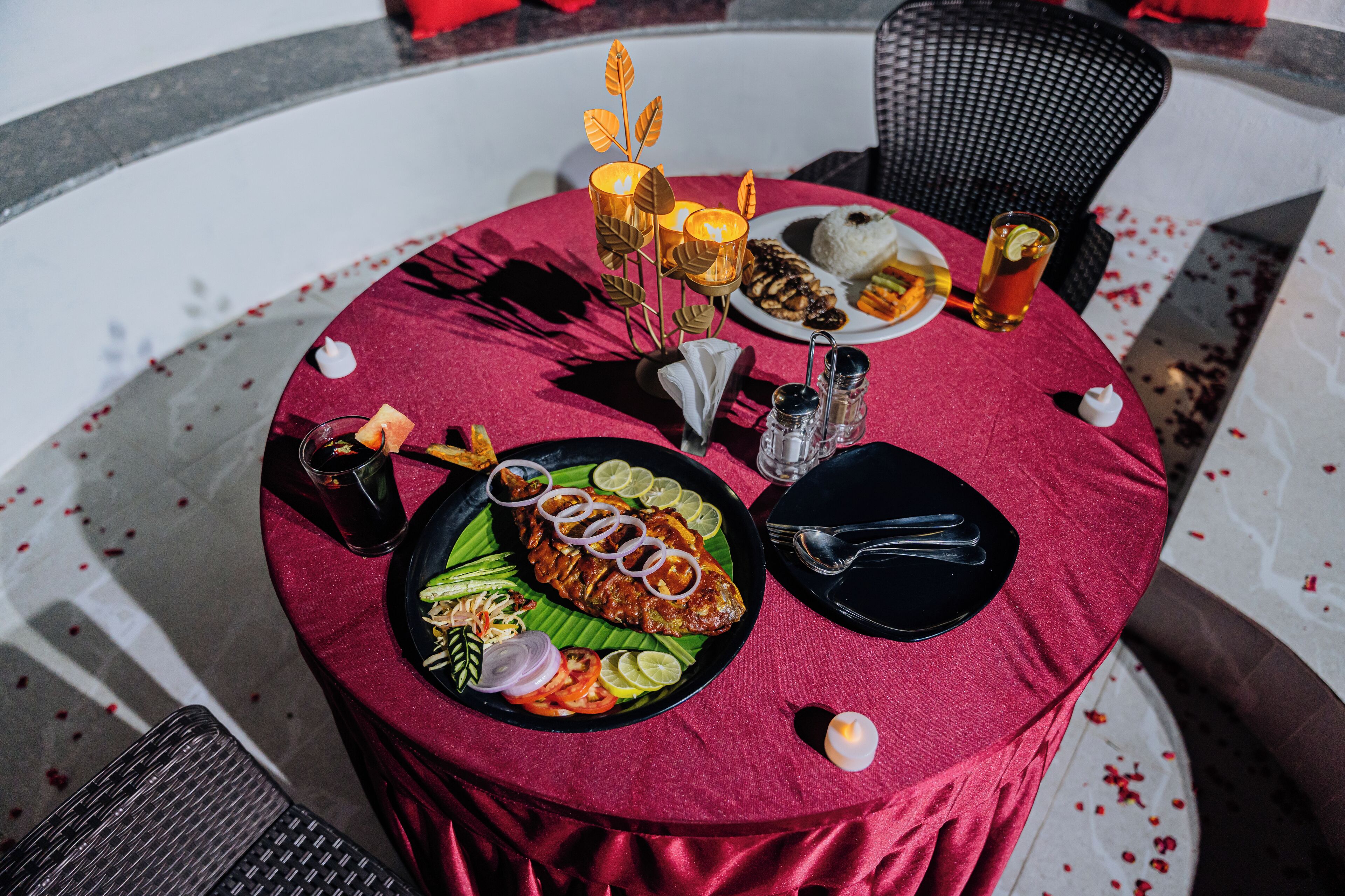 A round table with a red tablecloth, a plate of food, a glass of drink, and candles.