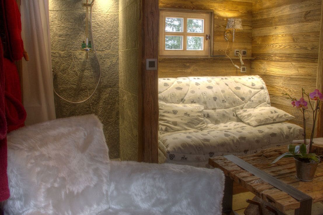 Quarto acolhedor em estilo cabana, com decoração em madeira, cama com roupa de cama estampada, área de chuveiro e uma pequena mesa com uma planta.