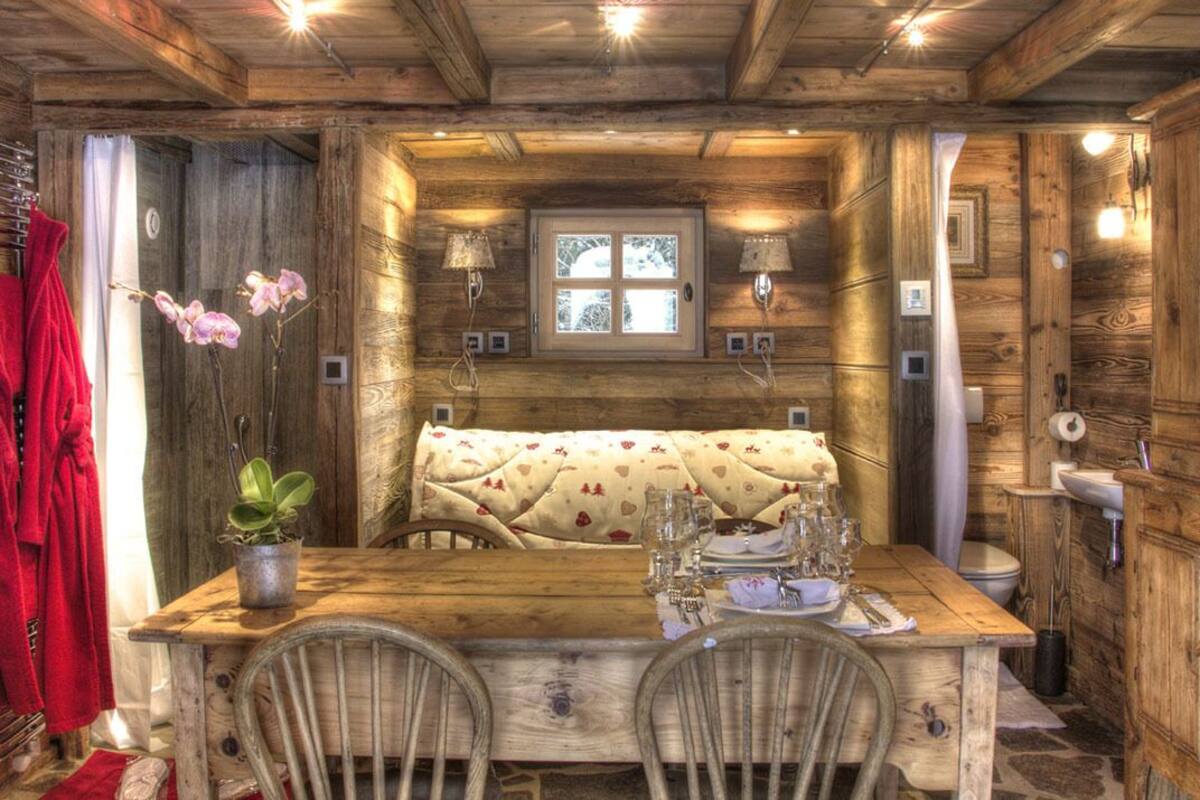 Interno rustico di una baita di legno con un tavolo da pranzo apparecchiato per due persone, una panca e una pianta in vaso.