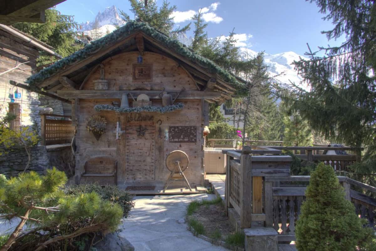 Un rustico chalet in legno con fondamenta in pietra, un balcone in legno e un piccolo giardino con pini.