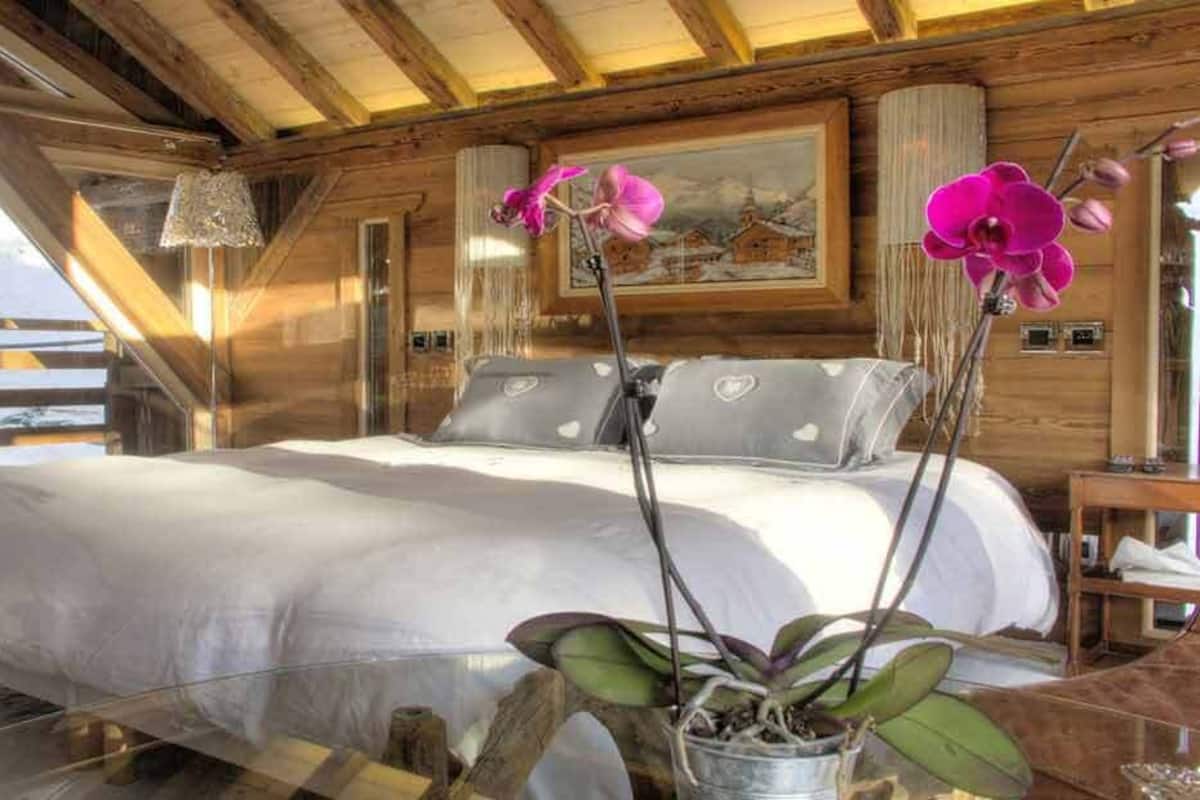Camera da letto di una baita in legno con un letto, cuscini, un comodino e una finestra vista su un paesaggio innevato.