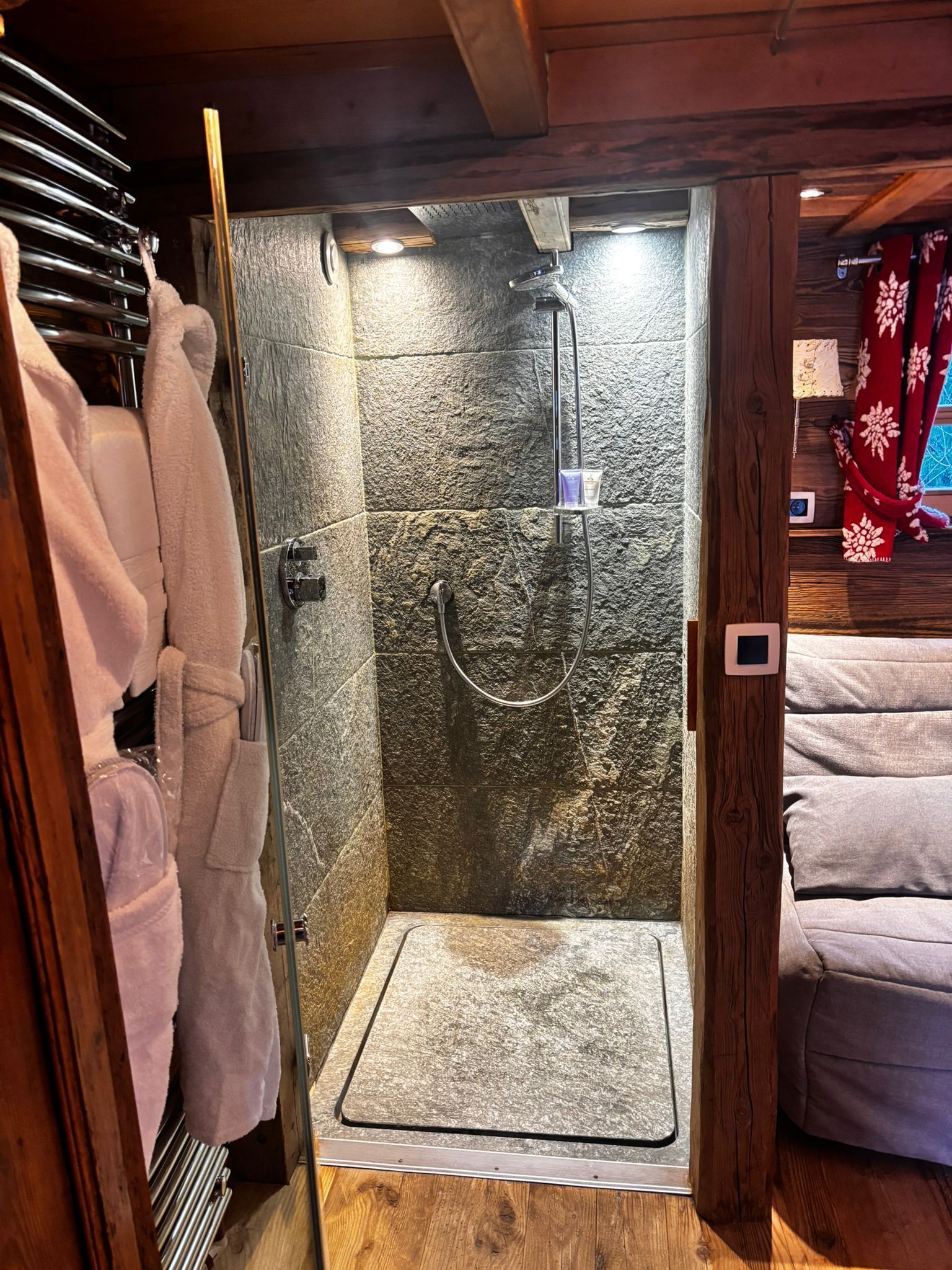 A shower with a stone wall, a glass door, and a wooden floor.