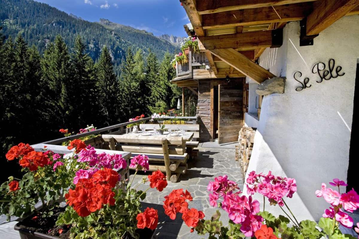 Un'area pranzo esterna in legno con tavolo in pietra apparecchiata per due, circondata da vivaci gerani.