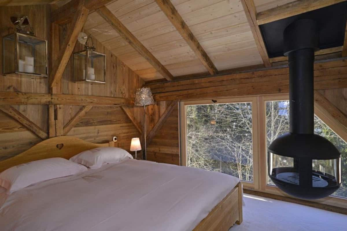 Camera da letto in una baita di legno con un letto grande, un lampadario e vista sugli alberi dalla finestra.