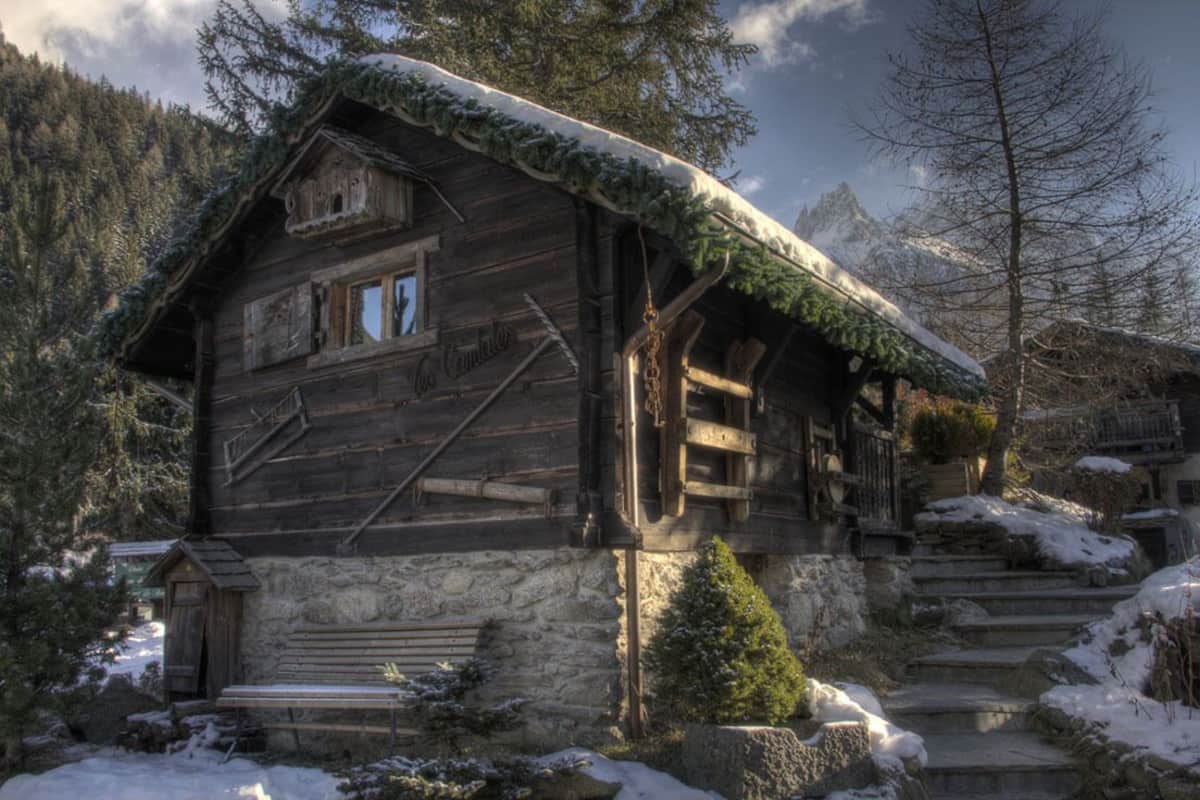 Una rustica baita di legno con fondamenta in pietra, circondata da alberi innevati e uno sfondo montuoso.