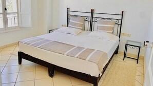 A neatly made bed with a striped bedspread and pillows, a small bedside table, a framed picture on the wall, and a window with a view of the outside. - Appart'hotel de charme  (Hammamet)